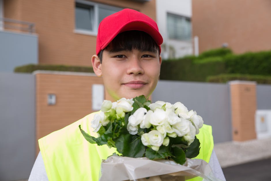 A young male flower delivery courier wearing a red cap and neon yellow vest holding a bouquet of fresh white roses with green leaves, wrapped in white paper, during daytime. The background features mo