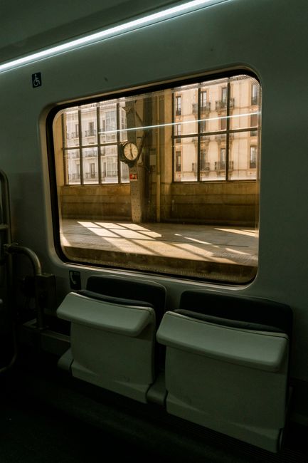 This image shows the interior of a train carriage with a large window revealing an urban scene outside. The window is framed by a sleek, light-colored interior panel. Just below the window, there are 