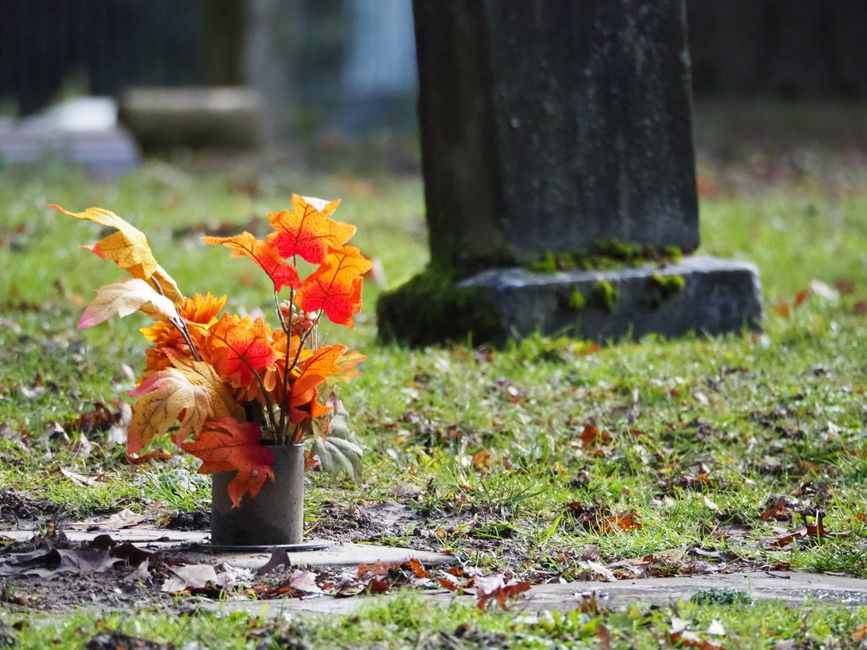 A bouquet of artificial autumn leaves in shades of orange, red, yellow, and cream arranged in a small gray vase, placed on a patch of grass near a gravestone in a cemetery setting. The leaves have a t