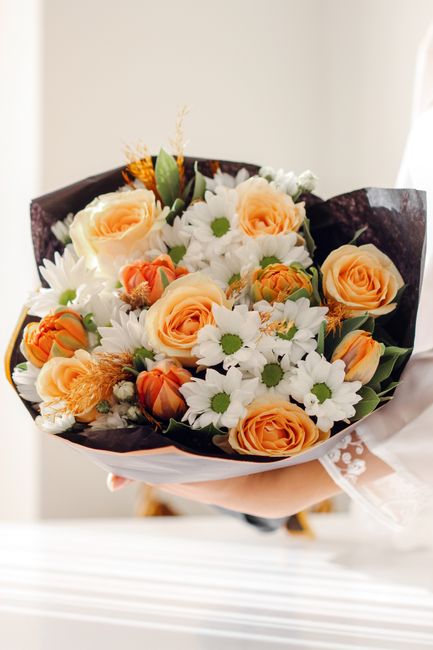 A bouquet of fresh flowers featuring peach-colored roses with fully opened petals, white daisy-like blossoms with yellow centers, and small orange-tinted buds. The flowers are arranged with green foli
