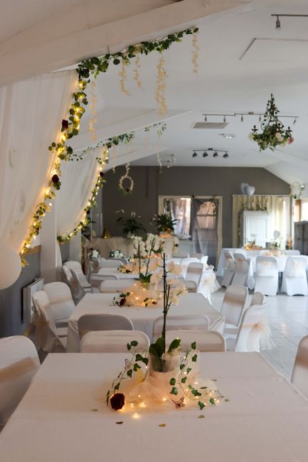 A spacious wedding reception room decorated with elegant floral arrangements and string lights. The tables are draped with white tablecloths and adorned with small vases holding white roses and greene
