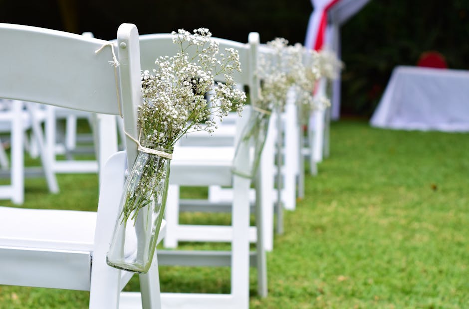 A row of white wooden chairs arranged outdoors on a grassy area, each adorned with a simple glass vase containing delicate white baby's breath flowers tied with natural twine. The arrangement creates 